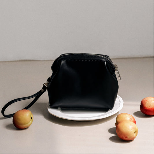 Labovarde Shell Pouch Bag in Black — Close-up of the leather bag with wrist strap on a white plate, with scattered Apples